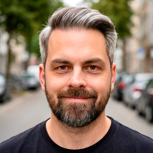Porträt eines lächelnden Mannes mit kurz geschnittenem grau meliertem Haar und Bart, der vor einer verschwommenen Straßenkulisse steht.