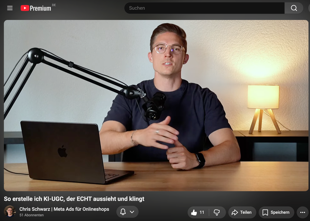 Ein Mann mit Brille sitzt an einem Holztisch vor einem Mikrofonarme, einem Laptop und einer kleinen Lampe auf einem Regal, während er in die Kamera spricht.